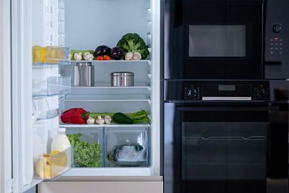 Бытовая техника на кухне: открытый холодильник с продуктами, микроволновая печь и духовой шкаф