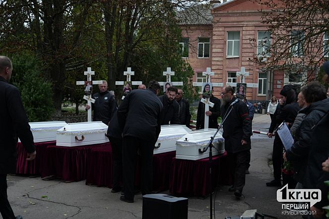 У селищі Радушне чоловіки встановлюють білі труни на підставки, позаду стоять люди з похоронними хрестами та фотографіями п'ятьох дітей, загиблих від російського удару.